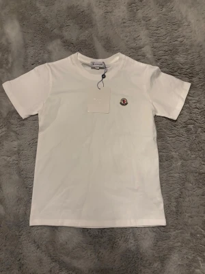 Vit Moncler t-shirt med logga - Snygg vit t-shirt från Moncler med klassisk rund hals och korta ärmar. På bröstet sitter Monclers ikoniska logga som en liten patch. Tillverkad i mjuk bomull för skön känsla och stilren look. Perfekt till jeans eller shorts. Passar dig som är ungefär 160-165cm.