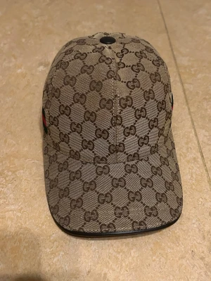 Gucci beige GG-mönstrad keps - Säljer en beige keps från Gucci med det klassiska GG-monogrammet i brunt över hela kepsen. Kepsen har böjd skärm och justerbart spänne bak. Materialet är canvas med läderdetaljer. Perfekt för dig som vill ha en lyxig och trendig accessoar.