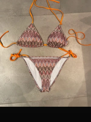 Mönstrad trekantsbikini med orange band - Säljee för högt pris eftersom osäker om jag vill sälja !!!! Köpte från vinted! Säljer en snygg trekantsbikini med zigzag-mönster i rosa, beige och brunt. Bikinin har knytband i stark orange färg både upptill och i sidorna på underdelen. Materialet är stretchigt och snabbtorkande, perfekt för stranden eller poolen.