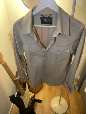 Beige overshirt från Oscar Jacobson - Stilren beige overshirt från Oscar Jacobson med klassisk krage, två bröstfickor med lock och knappar framtill. Tillverkad i ett mjukt bomullstyg och har långa ärmar med knapp vid manschetten. Perfekt för lager på lager och enkel att matcha.