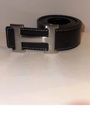 Mörkblått bälte  - Snyggt vändbart bälte i marinblått skinn med ett stort format H. Bältet har vita kontrastsömmar längs kanterna och klassisk design som sticker ut. Perfekt accessoar för att lyfta din outfit.