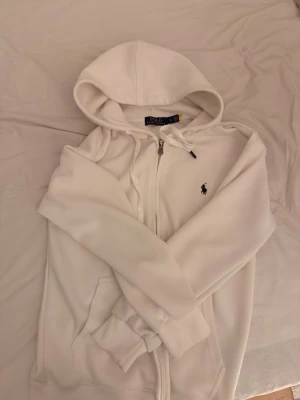 Vit hoodie från Polo Ralph Lauren - Vit hoodie med dragkedja från Polo Ralph Lauren. Klassisk design med huva, fickor framtill och den ikoniska lilla broderade loggan på bröstet. Mjuk bomullskänsla och ribbade muddar vid ärmslut och nederkant. Perfekt för en clean och stilren look.