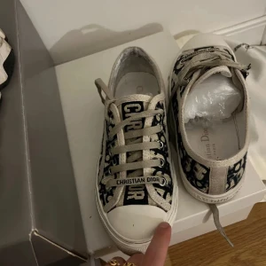 Christian Dior sneakers/Skor ”walk’N’Dior - Snygga sneakers/skor från Christian Dior med marinblått monogrammönster. Skorna har snörning, vit gummisula med Dior-logga och rund tå. Ovandelen är i canvas med tydliga Dior-detaljer och klassisk streetstil. Äktahetsbevis finns, samt dior box, och Dustbag  och de mörkblåa sko snörena. Det reducerade priset beror på den deffekten inget som märks av när man bär skorna, se sista bilden