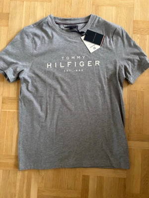 Grå t-shirt från Tommy Hilfiger - Snygg grå t-shirt från Tommy Hilfiger med klassisk logga tryckt framtill. Perfekt basplagg för en clean och stilren look. Kan gå ner i pris vid snabbt köp.