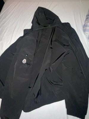 Svart vindjacka från Moncler med huva - Säljer en svart vindjacka från Moncler i storlek M. Jackan har huva, dragkedja framtill och en klassisk Moncler-logga på ärmen. Tillverkad i lätt polyester som gör den perfekt för blåsiga dagar. Insidan har ett coolt serietryck och jackan har en clean, stilren look. Men dragkedjan är sönder priset kan diskuteras.