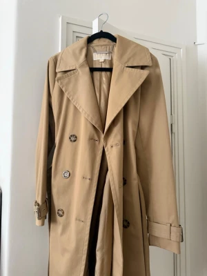 Beige trenchcoat från Michael Kors - Stilren beige trenchcoat från Michael Kors med klassisk dubbelknäppning, bred krage och bälte i ärmsluten. Jackan har en tidlös design och är tillverkad i ett slätt tyg som ger en snygg siluett. Perfekt för dig som vill ha en ikonisk och trendig look.