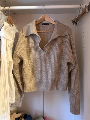 Beige stickad tröja med krage - Säljer en beige stickad tröja med bred krage och v-ringning. Tröjan har en avslappnad passform, ribbstickad struktur och långa ärmar. Perfekt för lager på lager och enkel att styla till olika outfits.