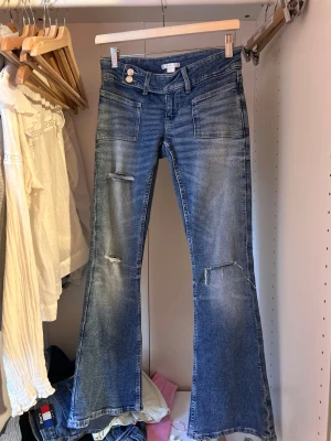 Blå slitna bootcut jeans Gina Tricot - Säljer ett par blå bootcut jeans från Gina Tricot med slitningar och hål på benen. Jeansen har hög midja, framfickor och en cool tvättad look. Perfekta för dig som gillar en avslappnad och trendig stil.