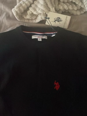 Svart tröja från U.S. Polo Assn - Klassisk svart tröja från U.S. Polo Assn med rund halsringning och röd broderad logga på bröstet. Tröjan har långa ärmar och en enkel, stilren design som passar till det mesta. Perfekt för dig som gillar en clean och tidlös look. Aldrig använd! Prislappen kvar. 