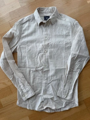Beige linneskjorta från H&M - Snygg beige skjorta från H&M i linne med klassisk krage och knappar framtill. Skjortan har lång ärm och en avslappnad regular fit som passar perfekt till både jeans och chinos. Enkel och stilren design som funkar året runt.