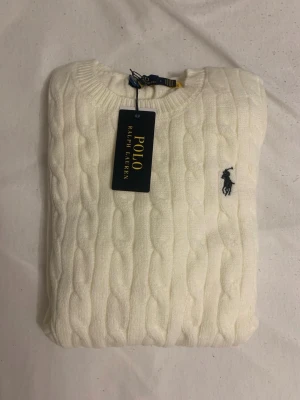 Vit kabelstickad tröja Polo Ralph Lauren - Kabelstickad vit tröja från Polo Ralph Lauren med rund halsringning och klassisk marinblå logga broderad på bröstet. Tröjan har ribbade muddar vid ärmslut och nederkant. Perfekt för dig som gillar stilrena och tidlösa plagg.mått: axelbredd 40cm. Längd 64cm