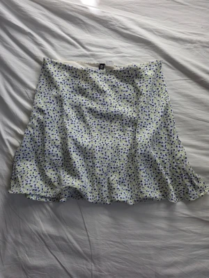 Vit blommig kjol från H&M - Supersöt kjol i väldigt bra skick från H&M i vitt med små blå och gröna blommor. Kjolen har en lätt utsvängd form. Enkel att matcha med både t-shirt och linne.
