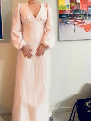 Ljus långklänning med puffärmar - Elegant ljus långklänning i satinliknande material med djup v-ringning, puffiga långärmar och öppen rygg med knytband. Klänningen har en mjuk, skimrande yta och en figurnära passform upptill som faller rakt ner. Den köptes förra året men har ej användes. Ny pris:1500kr