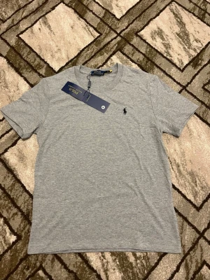 Grå t-shirt från Polo Ralph Lauren - Snygg grå t-shirt från Polo Ralph Lauren med klassisk passform och liten broderad logga på bröstet. Tillverkad i mjuk bomull och har rund halsringning samt korta ärmar. Perfekt basplagg för en clean och stilren look.