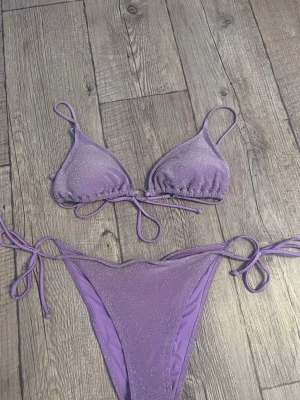 Lila glittrig bikini med knyt - Trendig lila bikini med glittrigt tyg och knyt i både topp och trosa. Triangelformad överdel och högt skurna trosor. Hör av dig om du har frågor🥰