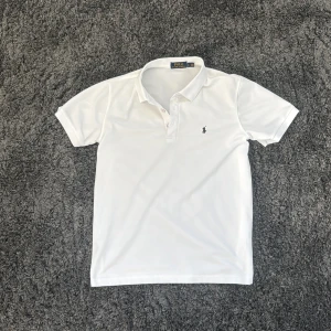 Vit pikétröja från Polo Ralph Lauren - Klassisk vit pikétröja från Polo Ralph Lauren med broderad svart logga på bröstet. Tröjan har korta ärmar, krage och knäppning framtill. Perfekt för en clean och stilren look, funkar året runt och är gjord i bomull för skön känsla.