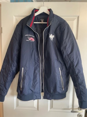 Marinblå bomberjacka Vinson Polo Club - Snygg marinblå bomberjacka från Vinson Polo Club med vita detaljer och broderade loggor på bröstet. Jackan har dragkedja framtill, två fickor med dragkedja och ribbade muddar. Insidan av kragen har en röd rand som ger extra stilpoäng.