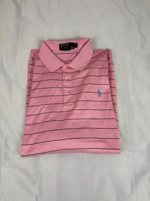 Rosa randig piké från Polo Ralph Lauren - Klassisk rosa pikétröja från Polo Ralph Lauren med tunna mörkblå ränder och broderad logga på bröstet. Tröjan har krage, knäppning med tre knappar och är tillverkad i mjuk bomull. Perfekt för en avslappnad men stilsäker look.