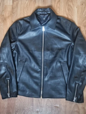 Svart skinnjacka från H&M - Snygg svart skinnjacka från H&M med raka linjer och klassisk krage. Jackan har silverfärgad dragkedja framtill och vid ärmsluten samt två snedställda fickor. Perfekt för dig som vill ha en stilren och tidlös look.