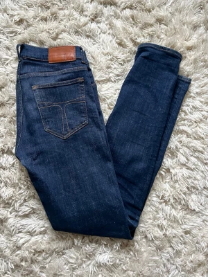 Mörkblå slim fit jeans från Tiger - Snygga mörkblå slim fit jeans från Tiger of sweden med klassiska bakfickor och kontrastsömmar. Jeansen har en läderpatch med logga bak i midjan och är tillverkade i ett stretchigt denimtyg för skön passform.