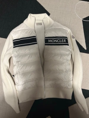Vit pufferjacka från Moncler - Snygg vit pufferjacka från Moncler med stickade ärmar och hög krage. Jackan har svarta och vita ränder över bröstet samt Moncler-logga på vänster ärm. Dragkedja framtill och quiltad front ger en modern look. Helt oanvänd!!!!  A kopia