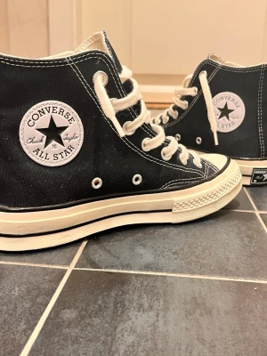 Converse Chuck Taylor All Star 70s - Klassiska svarta Converse Chuck Taylor All Star 70s med högt skaft, vita kontrastsömmar och vita snören. Skorna har en vit gummisula med svart rand och ikonisk logga på sidan. Ovandelen är i slitstark canvas och sulan i gummi.