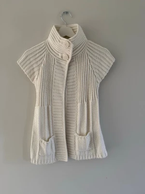 Vit stickad kofta - Säljer en krämvit stickad kofta från Amisu med korta ärmar, hög krage och två stora knappar framtill. Bra skick då den inte kommit till användning och i storlek m.