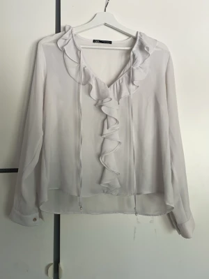 Vit volangblus från Zara - Supersöt vit blus från Zara med långa ärmar och volangdetaljer längs den djupa v-ringningen. Blusen är gjord i ett tunt, lätt material som ger ett flowy intryck. Perfekt för dig som gillar romantiska detaljer och en luftig känsla.