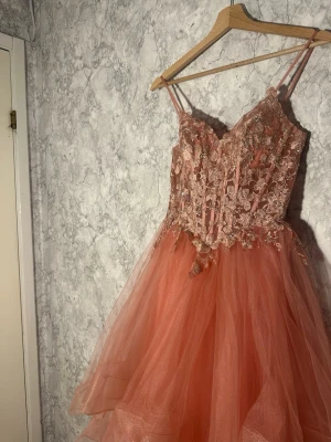 Rosa långklänning med spetsdetaljer - Romantisk rosa långklänning med tunna axelband och vacker spets upptill. Klänningen har volangkjol i flera lager och snörning i ryggen för en justerbar passform. Perfekt för speciella tillfällen där du vill känna dig extra fin.