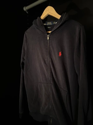 Mörk blå zip från Polo Ralph Lauren - Mörk blå hoodie med dragkedja från Polo Ralph Lauren. Klassisk design med röd broderad logga på bröstet, huva och kängurufickor framtill. Tillverkad i mjukt material som är skönt att ha på sig.