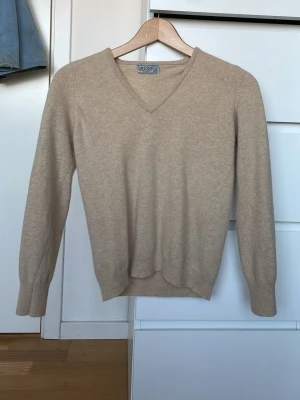 Beige V-ringad kashmirtröja - Snygg beige stickad tröja i mjuk kashmir. Tröjan har V-ringning, långa ärmar och ribbade muddar vid ärmslut och nederkant. Perfekt för lager på lager och enkel att matcha med olika stilar.