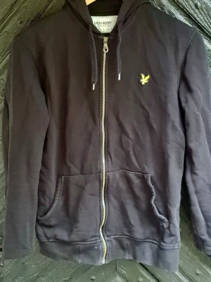 Svart hoodie från Lyle & Scott - Svart zip-hoodie från Lyle & Scott med gul broderad logga på bröstet. Hoodien har dragkedja, två stora fickor fram och justerbar huva med snören. Tillverkad i mjukt bomullsmaterial och har ribbade muddar vid ärmslut och nederkant.