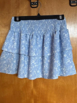 Blå volangkjol med blommönster - Supersöt blå kjol med vita småblommor och bred smock i midjan. Kjolen har dubbla volanger som ger en flowy och lekfull look. Perfekt för dig som gillar en feminin och trendig stil. Materialet ser ut att vara mjukt och lätt, perfekt för varma dagar.           Storlek 158/164