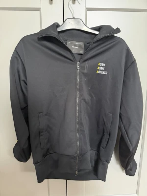 Svart zip hoodie Axel Arigato - Snygg svart hoodie från Axel Arigato med dragkedja framtill och två fickor. På bröstet finns ett broderat tryck i vitt och gult med texten 'ADEN ASME ARIGATO'. Perfekt för en avslappnad och trendig stil.