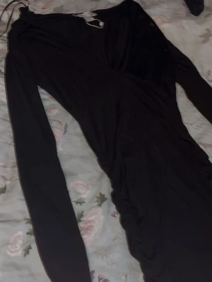 Svart långärmad omlottklänning - Säljer en svart långärmad klänning med omlottdesign och rynkade detaljer längs sidorna. Klänningen har en figurnära passform och är perfekt för dig som gillar en stilren och elegant look. Materialet känns mjukt och stretchigt.