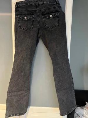 Grå bootcut jeans med slitning - Säljer ett par grå bootcut jeans med lätt slitning och klassiska fem fickor. Jeansen har en något utsvängd passform nedtill och normal midja. Perfekta för dig som gillar en avslappnad men trendig look.