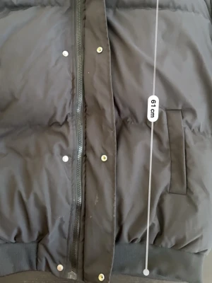 Svart pufferjacka med dragkedja - Säljer en snygg och bekväm svart pufferjacka – perfekt för kalla dagar. Jackan är både varm och lätt att bära, vilket gör den idealisk för vardagligt bruk.  Detaljer: 	•	Dragkedja framtill med tryckknappar 	•	Två snedställda fickor 	•	Ribbade muddar nedtill 	•	Bekväm och isolerande passform  Längd: ca 61 cm  En perfekt jacka för dig som vill hålla värmen utan att kompromissa med stilen.  💬 Hör av dig vid intresse!.