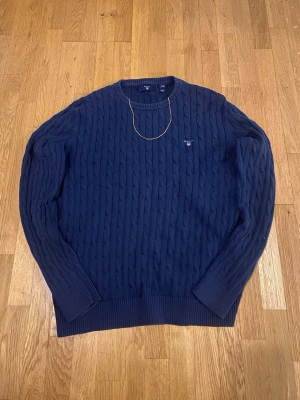 Marinblå stickad gant tröja  - Säljer en klassisk och stilren kabelstickad tröja från GANT i mörkblå färg. Perfekt för både vardag och lite mer uppklädda tillfällen.  ✨ Detaljer: 	•	Färg: Mörkblå 	•	Modell: Kabelstickad 	•	Passform: Bekväm och något avslappnad 	•	Material: Mjuk och skön kvalitet 	•	Broderad GANT-logga på bröstet.                                 Litet hål på vänstra armbågen ( se sista bilden) och så ärlappen med  loggan brevid storleken lös på vänstra sidan