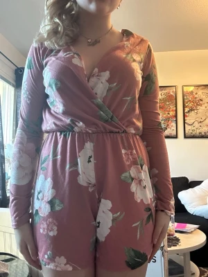 Rosa blommig långärmad jumpsuit - Supersöt rosa jumpsuit med vita och gröna blommor. Långärmad modell med omlott-ringning och resår i midjan för en skön passform. Kort byxdel och mjukt, stretchigt tyg som känns bekvämt hela dagen. Perfekt för dig som gillar en romantisk och trendig stil.