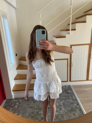 Vit axelbandslös kort klänning - Söt vit axelbandslös kort klänning med smockad överdel och volangig kjol. Klänningen har broderade spetsdetaljer och är perfekt för varma dagar. Lätt och luftig modell som ger en romantisk känsla.