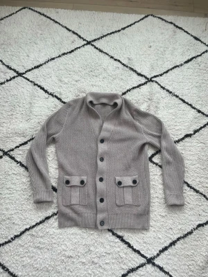 Kofta - Säljer en grå kofta med v-ringning och svarta knappar framtill. Koftan har två stora fickor med dekorativa lock och knappar samt ribbade muddar vid ärmslut och nederkant. Perfekt för lager på lager och en chill look.