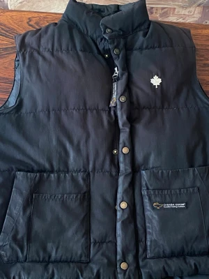 Svart dunväst från Canada Goose - Snygg svart dunväst från Canada Goose med hög krage, tryckknappar och dragkedja framtill. Västen har två stora framfickor och en vit lönnlogga på bröstet. Perfekt för lager-på-lager och streetwear-stil.