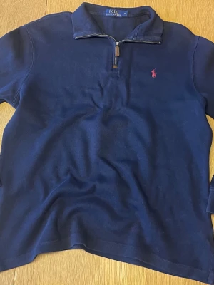 Marinblå halvzip från Polo Ralph Lauren - Klassisk marinblå tröja med halv dragkedja från Polo Ralph Lauren. Tröjan har en liten rosa logga broderad på bröstet och är tillverkad i mjukt stickat material. Perfekt till jeans eller chinos för en clean look.  70 cm längd, 58 cm axel till axel