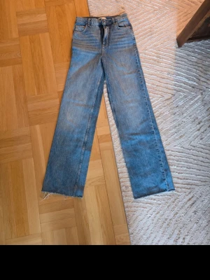 Blå raka jeans med rå kant gina tricot  - Säljer ett par blå raka jeans med hög midja och rå, fransig kant längst ner på benen. Klassisk femficksmodell i slitstarkt denimtyg. Perfekta för en avslappnad och trendig look.