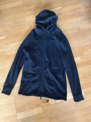 Svart hoodie från Tiger of Sweden - Säljer en svart hoodie från Tiger of Sweden Jeans med dragkedja, huva med dragsko och två stora fickor framtill, i längre modell. Tröjan har rå kant nertill för en avslappnad look och är gjord i mjukt material. Perfekt för dig som gillar streetwear.