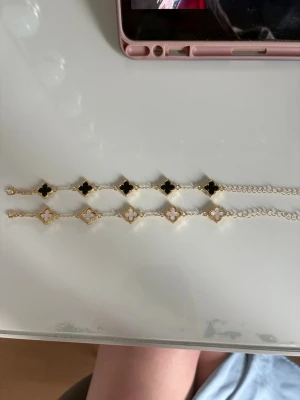 Guldarmband med clover-detaljer - Två eleganta armband i guldton med clover-formade hängen. Ett armband har svarta detaljer och det andra har vita pärlliknande inslag. Båda har tunn kedja och stilren design som ger en lyxig känsla.