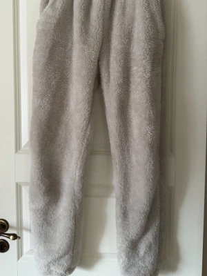 Grå fluffiga loungewear-byxor - Supermjuka och fluffiga loungewear-byxor i ljusgrå färg. Perfekta för att chilla hemma, med resår i midjan och avslappnad passform. Tillverkade i ett mysigt teddy-liknande material som känns extra skönt mot huden.