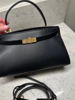 Svart handväska med gulddetaljer  - Stilren svart handväska i slätt läder från Salvatore Ferragamo. Väskan har ett elegant guldfärgat spänne framtill, matchande gulddetaljer och ett avtagbart axelband. Klassisk form med topphandtag och minimalistisk design.