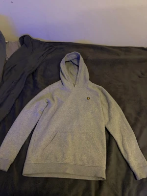 Grå hoodie från Lyle & Scott - Snygg grå hoodie från Lyle & Scott med klassisk gul logga på bröstet. Hoodien har huva, känguruficka framtill och ribbade muddar vid ärmar och nederkant. Tillverkad i mjukt bomullsmaterial som är skönt att bära.