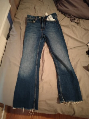 Blå bootcut jeans med rå kant - Säljer ett par blå bootcut jeans med rå, fransig kant nedtill. Jeansen har klassisk femficksmodell och en snygg tvätt med lätt slitning på låren. Perfekta för dig som gillar en avslappnad och trendig stil.